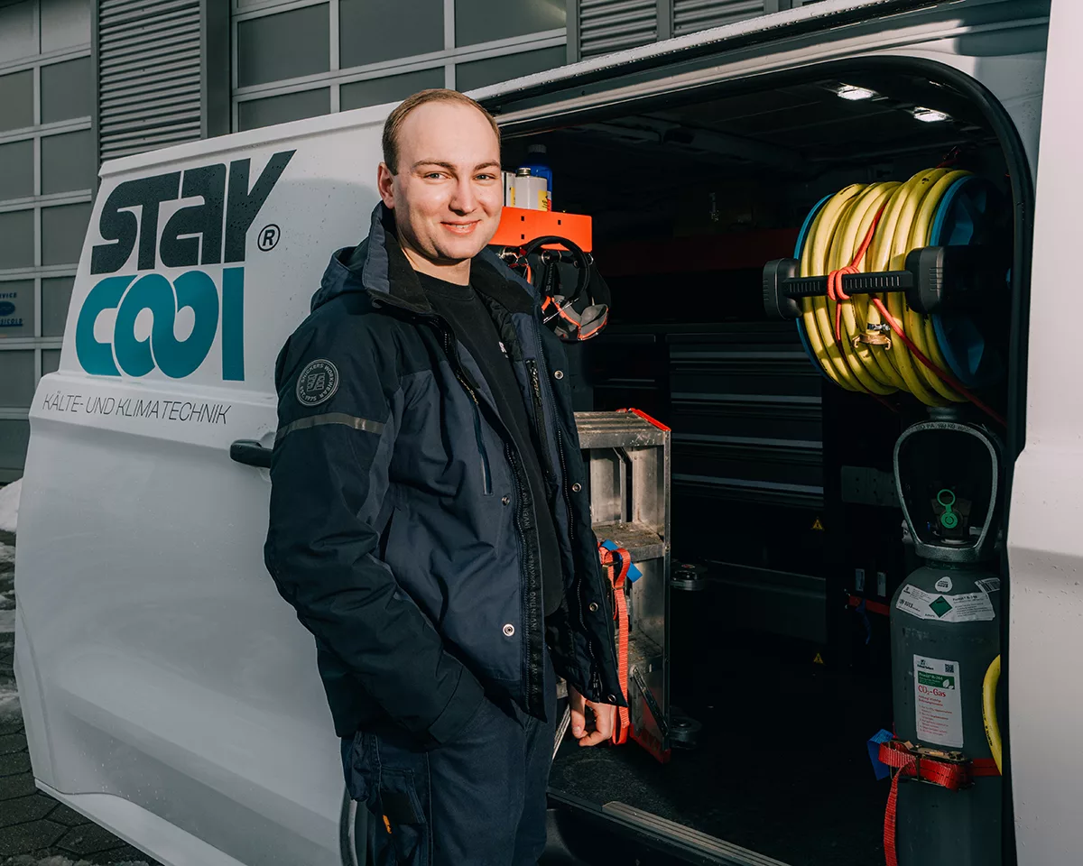 Ein Mitarbeiter der Kälte- und Klimatechnikfirma „Stay Cool” steht lächelnd vor dem Firmentransporter und blickt in die Kamera.