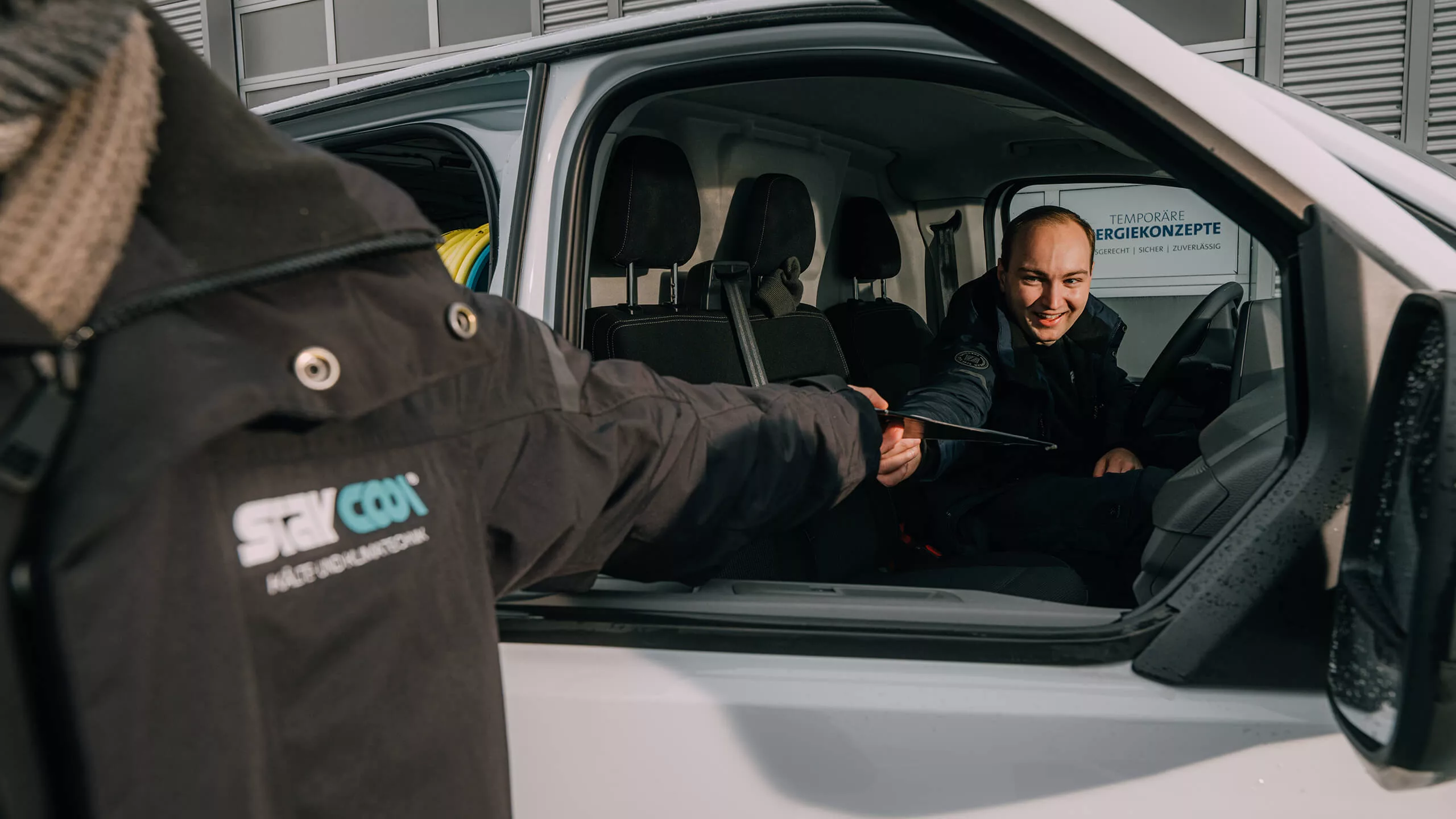 Ein Mitarbeiter der Kälte- und Klimatechnikfirma „Stay Cool” sitzt lächelnd in einem Firmentransporter gibt seinem Kollegen etwas entgegen.