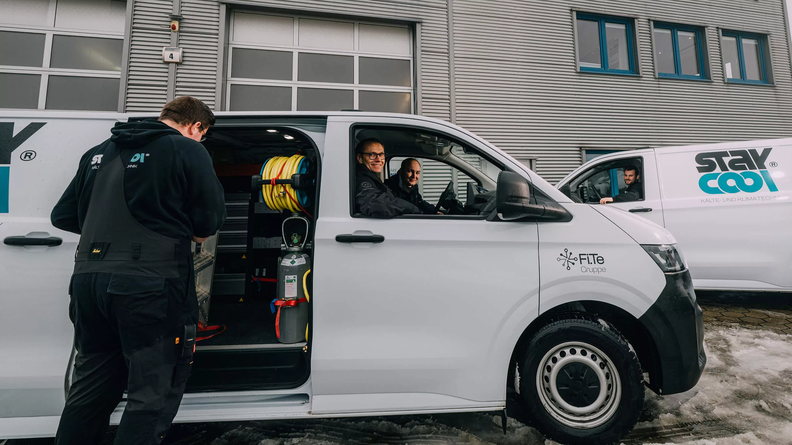 Ein Mitarbeiter von Stay Cool befindet sich vor der Werkstatt bei einem der Firmentransporter. Er räumt Werkzeug in den Transporter ein. Währenddessen sitzt einer der Mitarbeiter im Transporter am Lenkrad, der andere neben ihm auf dem Beifahrersitz. Im Hintergrund ist noch ein weiterer Transporter zu sehen, in dem ein Mitarbeiter sitzt.